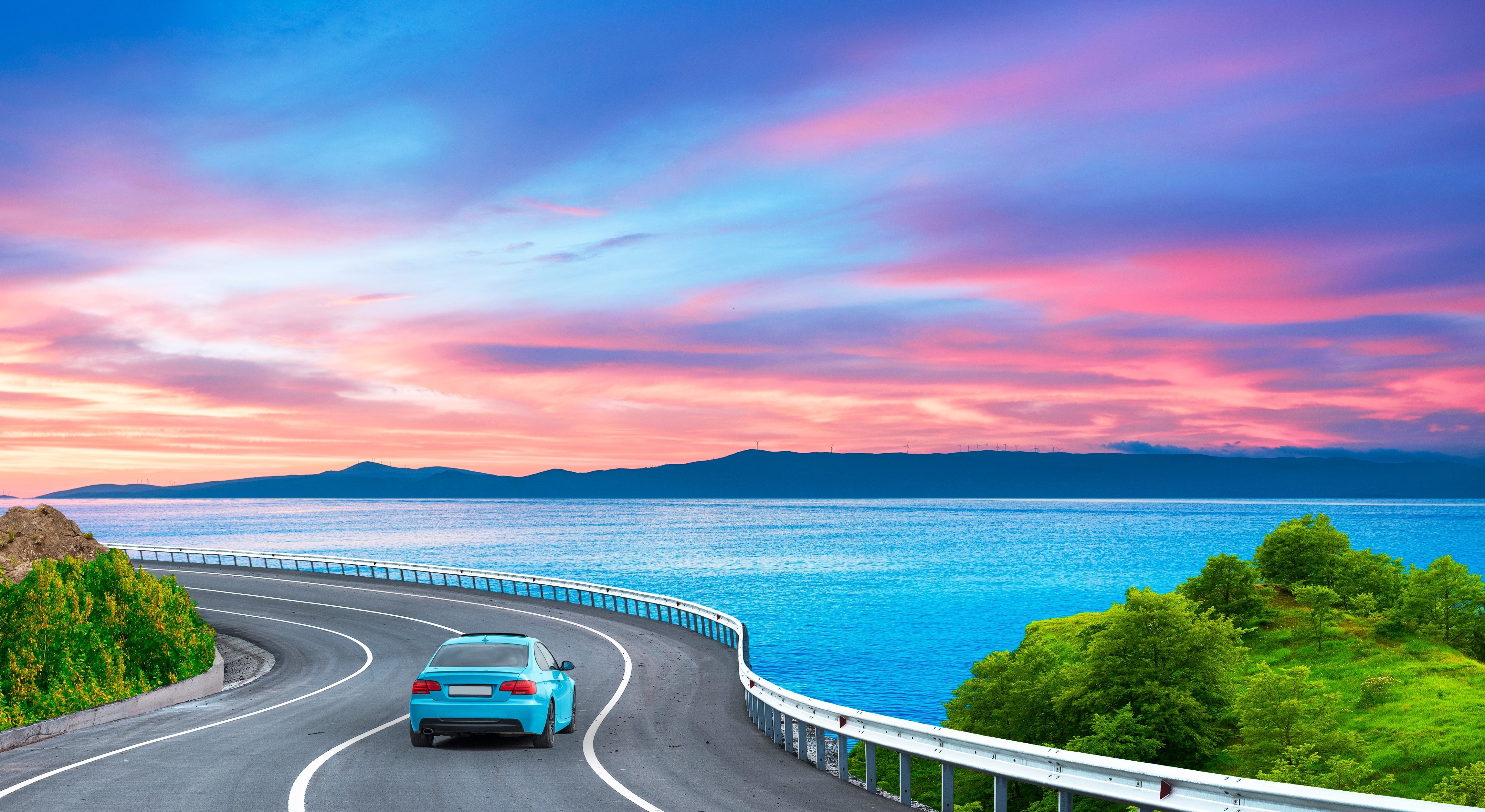 blue car driving along a coastal road towards the sunset